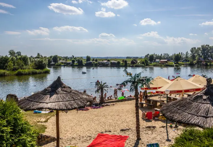 Słoneczny dzień na plaży w Kuźni Raciborskiej. Ludzie odpoczywają pod parasolami, kąpią się w jeziorze i pływają łódkami.