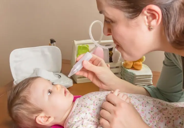 Mama pomaga noworodkowi z katarem, używając aspiratora. Dziecko leży na różowym ręczniku, patrząc na mamę.