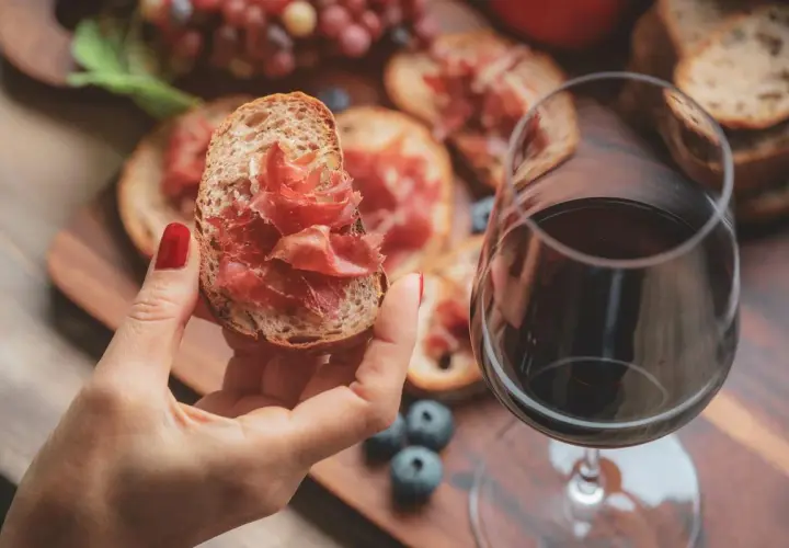 Une main tient une tartine garnie de jambon cru, à côté d'un verre de vin rouge. Parfait pour un apéro dinatoire.