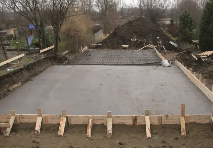 Plac budowy z wylana płytą betonową, przygotowywaną pod fundamenty. Widać zbrojenie i szalunki, wskazujące na różne klasy ekspozycji betonu.
