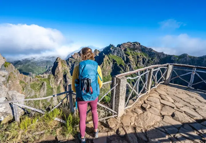 A hiker with a blue backpack enjoys a breathtaking view during a Madeira hike. Jagged, green mountains rise under a bright blue sky.