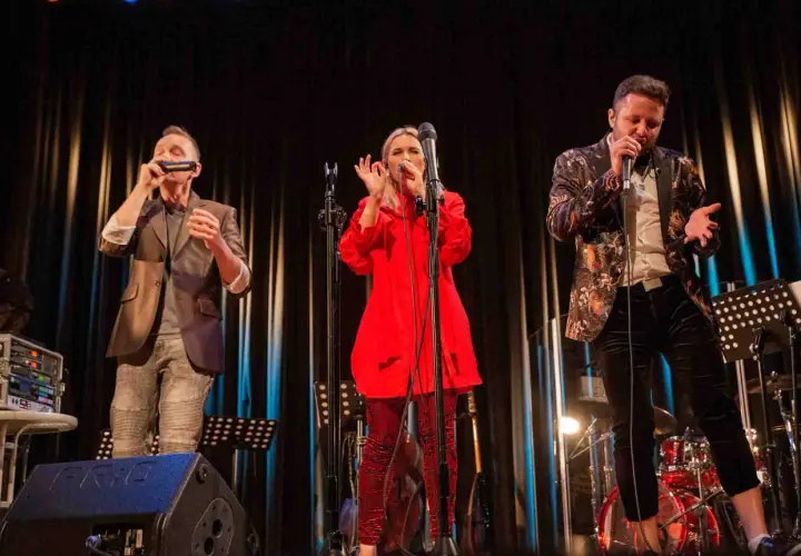 Trzech wokalistów na scenie podczas warsztatów wokalnych. Kobieta w czerwieni, mężczyzna w marynarce z wzorami i mężczyzna grający na harmonijce.