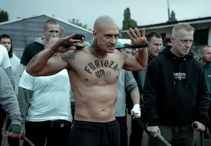 Wytatuowany mężczyzna z napisem "Furioza" na klatce piersiowej, otoczony przez grupę mężczyzn, przygotowuje się do konfrontacji.
