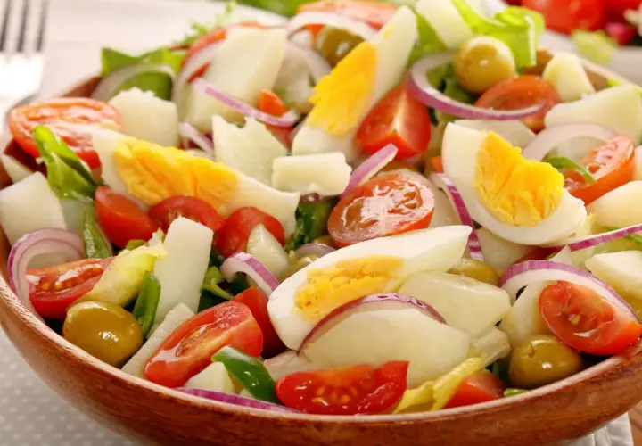 Deliciosa ensalada de patata con huevo duro, tomate, cebolla morada y aceitunas verdes en un cuenco de madera.