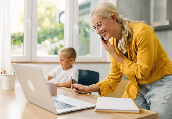Uśmiechnięta kobieta rozmawia przez telefon, pracując przy laptopie, podczas gdy jej syn rysuje obok. Odpowiedź na pytanie: czy na urlopie wychowawczym można pracować.