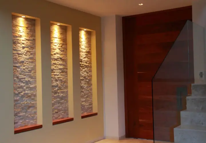 Tre nicchie in cartongesso illuminate da faretti, con rivestimento in pietra, creano un punto focale moderno accanto a una porta in legno e una scala con parapetto in vetro.