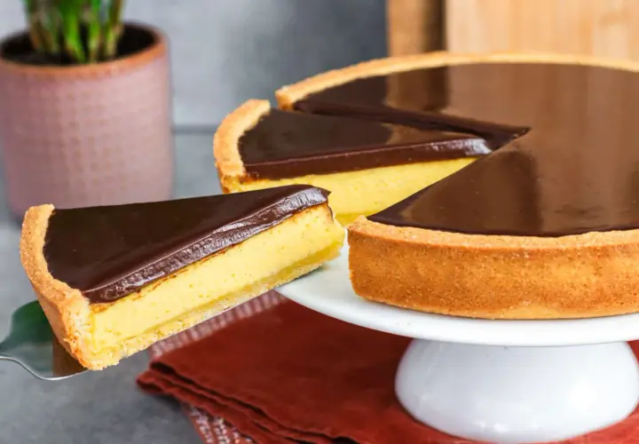 Una fetta di torta Susanna, con crema gialla e copertura al cioccolato fondente, viene sollevata da una tortiera su un'alzata bianca.