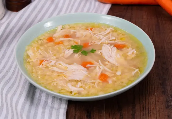 Un confortante brodo di pollo con pezzi di pollo sfilacciato, carote e un rametto di prezzemolo.