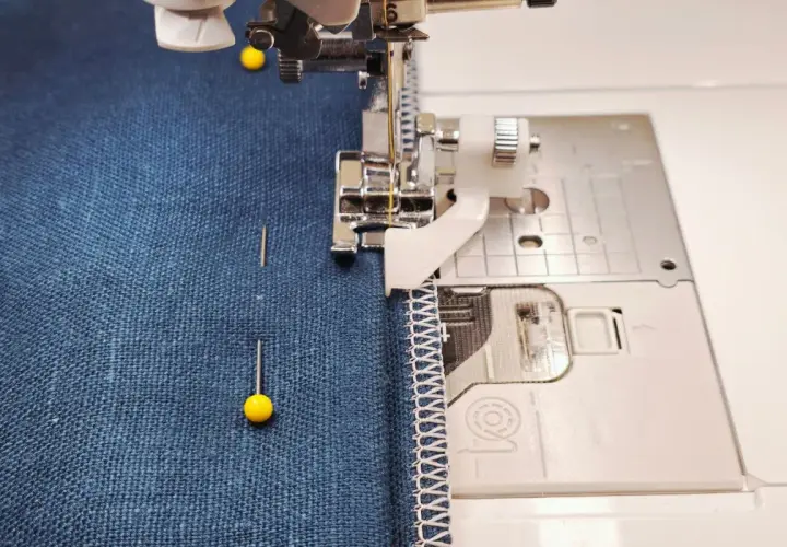 A sewing machine with a blind stitch foot is about to hem dark blue fabric. Pins hold the fabric in place.