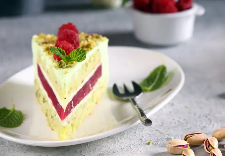 Pyszny tort pistacjowo malinowy z malinami i miętą na talerzu. Obok widoczne są pistacje.