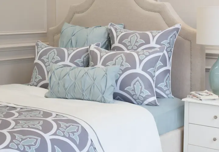 A bed with a tufted headboard, featuring large square pillows with a grey and white floral pattern, and smaller light blue accent pillows.