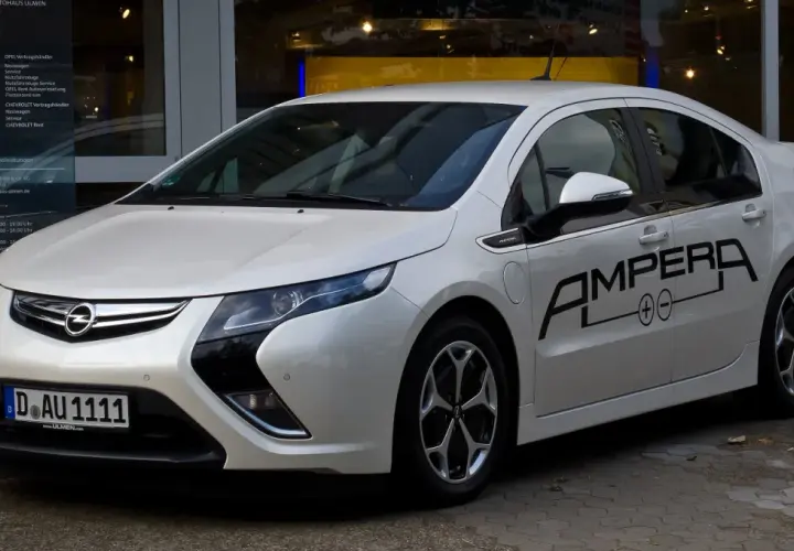 Biała Opel Ampera z czarnym napisem "AMPERA" na drzwiach, zaparkowana przed salonem samochodowym.