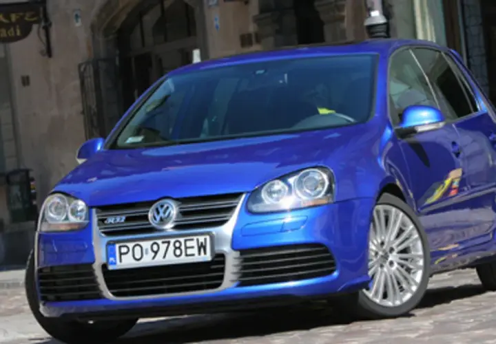 Błękitny Golf R32 z felgami ze stopów lekkich, zaparkowany na brukowanej uliczce przed kawiarnią.