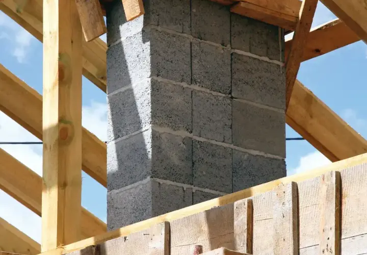 Budowa komina z bloczków betonowych, widoczne drewniane elementy konstrukcji dachu. Zbrojenie pod komin jest kluczowe dla jego stabilności.