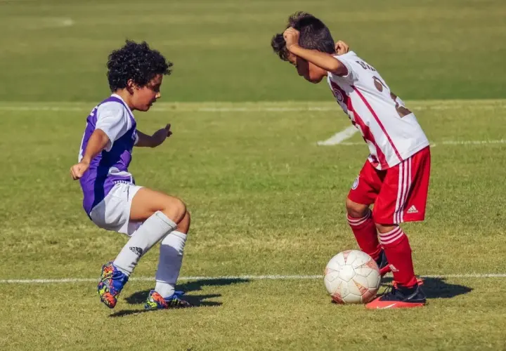 Due ragazzini si sfidano su un campo da calcio in erba, uno in maglia viola, l'altro in maglia bianca e rossa, con un pallone ai loro piedi.