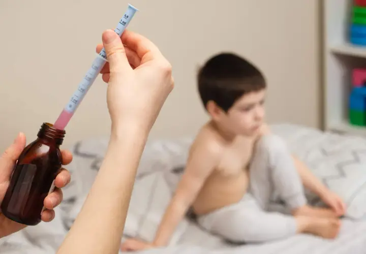 Mano preparando jeringa con ibuprofeno para un bebé. Niño en cama al fondo.