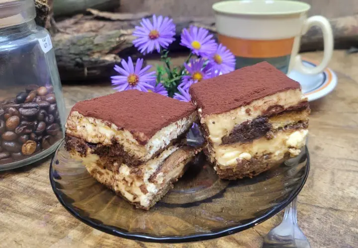 Dwa kawałki tiramisu, idealne ciasto do kawy, obok słoika z ziarnami kawy i fioletowych kwiatów.