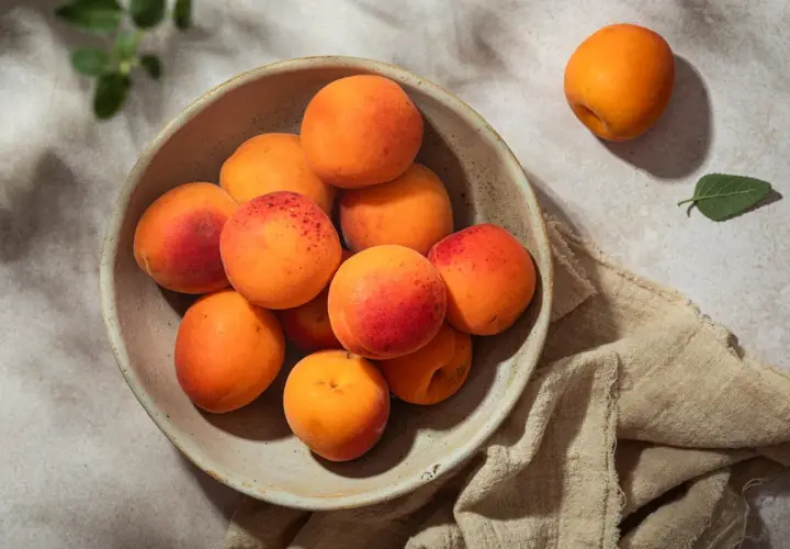 Un bol rempli d'abricots juteux, annonçant la belle **période abricot**. Un fruit isolé repose à côté, sur un tissu beige.