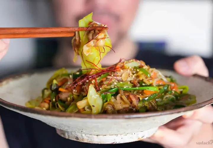 A person uses chopsticks to lift a bite of a colorful Japanese stir fry, featuring cabbage, carrots, and peppers, from a rustic bowl.