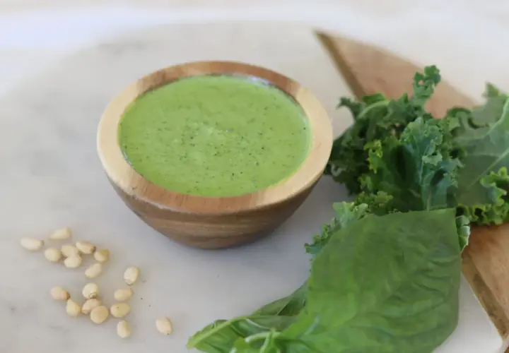 Un cuenco de madera contiene un vibrante pesto de kale, acompañado de hojas frescas de kale, albahaca y piñones.