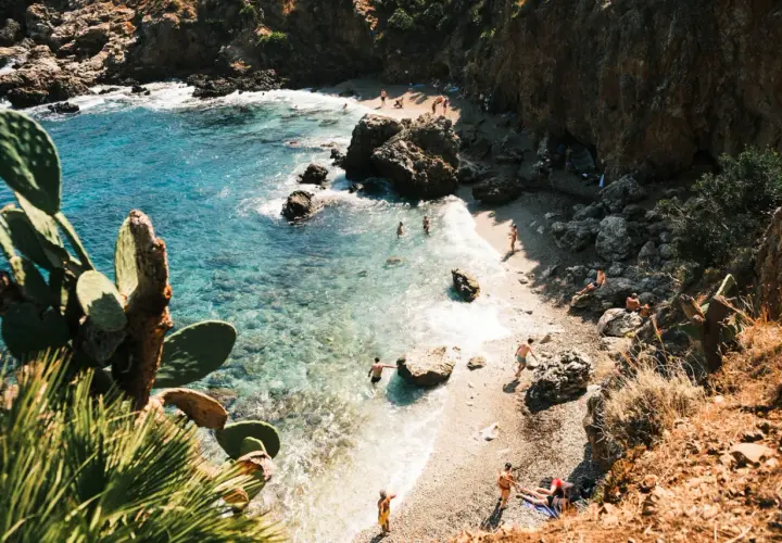 A secluded beach in Zingaro, Sicily, with crystal-clear turquoise water, rocky cliffs, and sunbathers enjoying the Mediterranean.