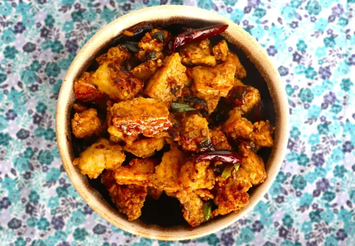 Crispy, golden-brown pieces of ice tofu are coated in a savory sauce with chili peppers and green onions, served in a bowl on a floral background.
