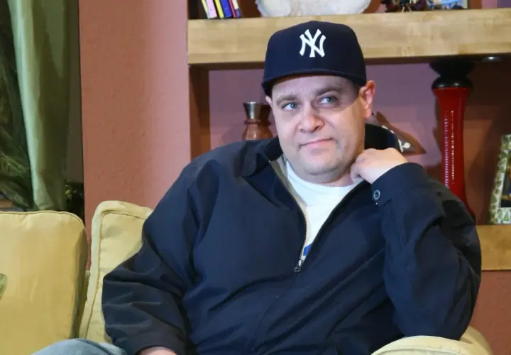 Mężczyzna w czapce z daszkiem Yankees, w wieku, który można by określić jako dojrzały, siedzi na kanapie.