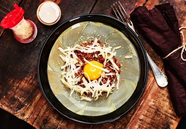 En läcker koreansk råbiff med en gul äggula i mitten, toppad med strimlad rättika och sesamfrön. Serveras på en vacker tallrik med en mörk servett och gaffel.