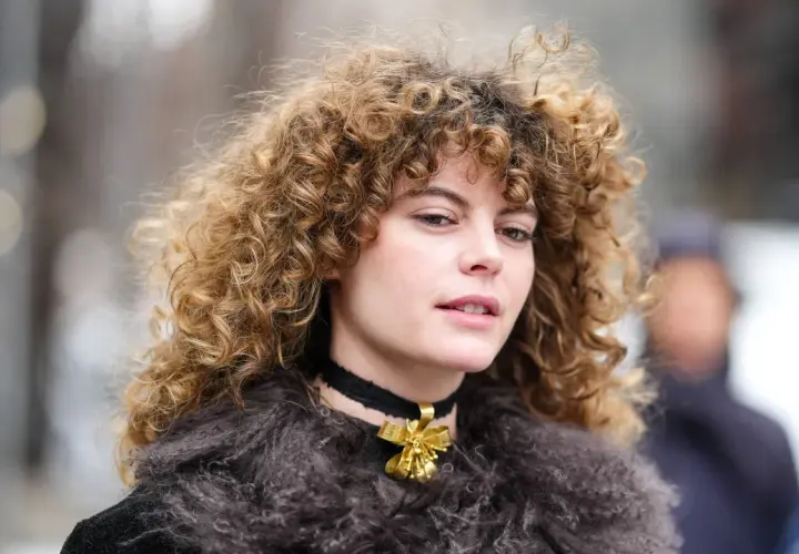 Eine Frau mit voluminösen, krausen Haaren trägt einen Pelzmantel und eine Halskette mit einer goldenen Schleife.