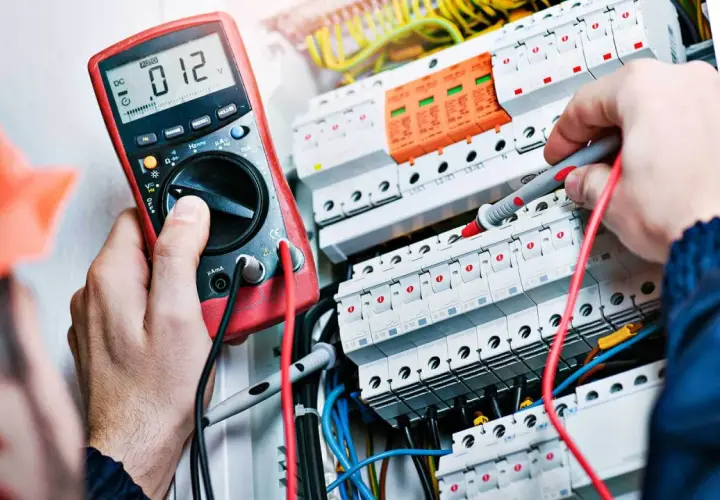 Elektryk w pomarańczowym kasku sprawdza instalację multimetrem. To pierwszy krok, jak zostać elektrykiem.