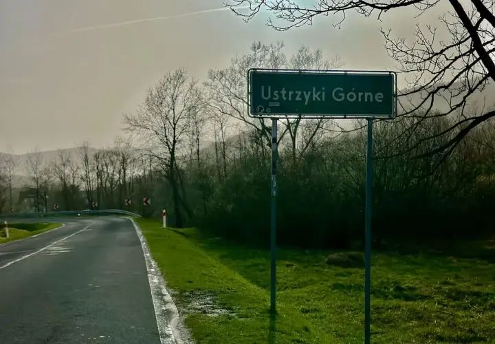 Znak drogowy z napisem "Ustrzyki Górne" na tle mokrej drogi i drzew.