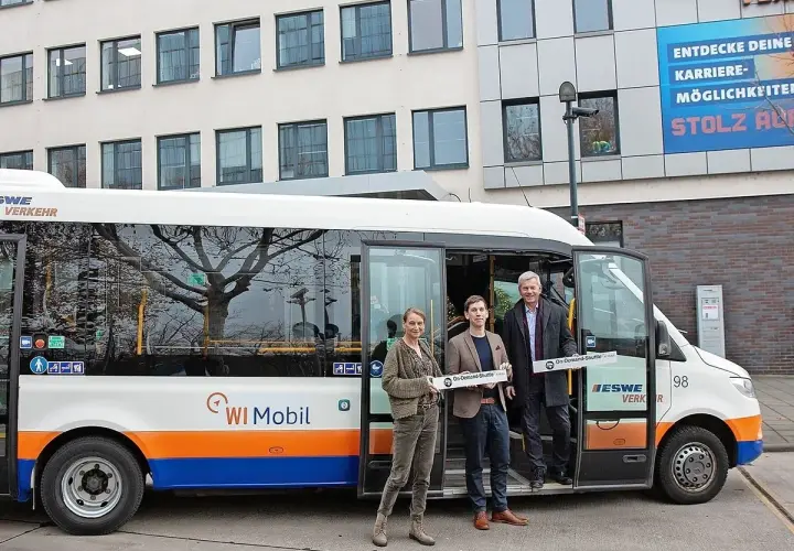 Drei Personen stehen vor einem weißen Kleinbus mit der Aufschrift "WI Mobil". Sie halten Schilder mit der Aufschrift "On-Demand-Shuttle".