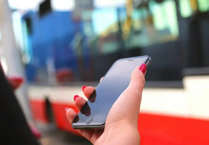 Dłoń z czerwonymi paznokciami trzyma smartfon, na którym można kupić bilety ZTM Warszawa. W tle widać autobus.