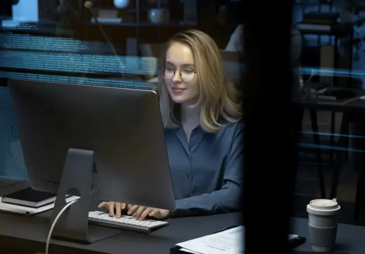Młoda kobieta w okularach pracuje przy komputerze, na ekranie widać kod. To przyszłość cyberbezpieczeństwa, studia w tej dziedzinie otwierają wiele możliwości.