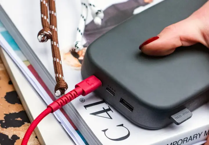 Dłoń z czerwonym lakierem podłącza czerwony kabel do powerbanku, zapewniając szybkie ładowanie.