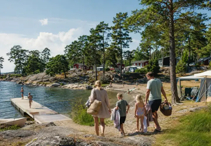Familj på väg mot vattnet för en härlig semester med bebis i Sverige. Barnen springer glatt mot bryggan där andra badar.