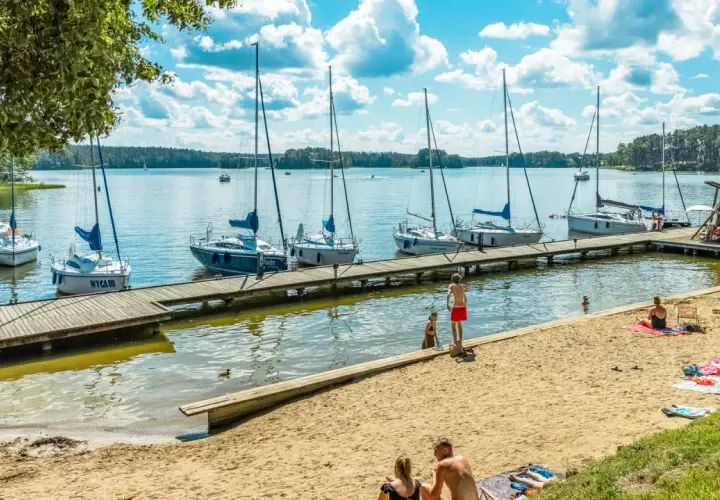 Słoneczne **jezioro z plażą**, gdzie cumują żaglówki. Ludzie odpoczywają na piasku i kąpią się w wodzie.