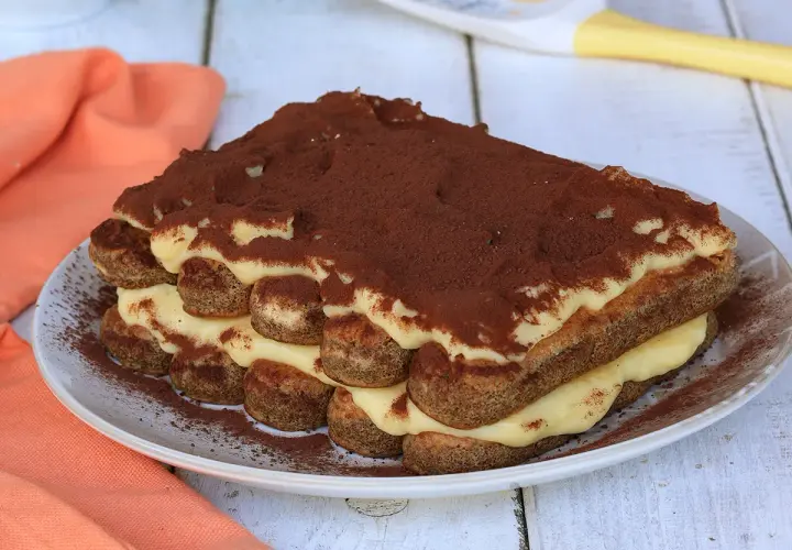 Tiramisù con crema pasticcera, strati di savoiardi e cacao in polvere su un piatto bianco, con un tovagliolo arancione sullo sfondo.