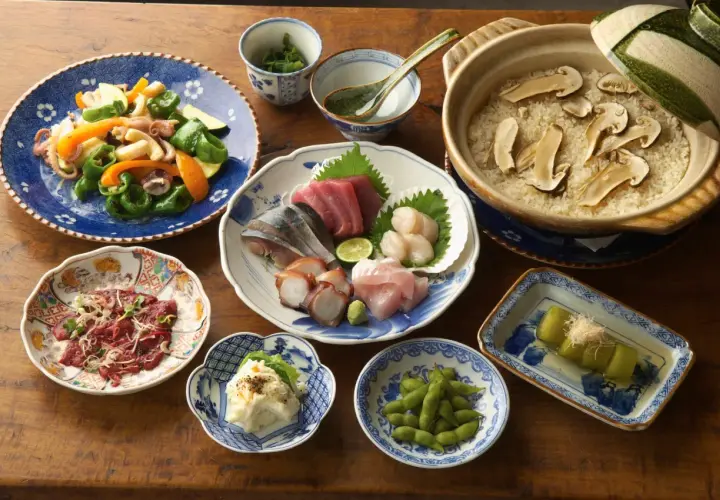 A spread of Japanese side dishes, including sashimi, edamame, potato salad, and a vegetable stir-fry, complements a pot of rice with mushrooms.