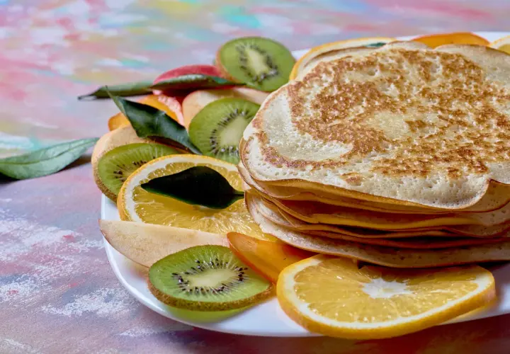 Stos naleśników z budyniem, otoczony plasterkami kiwi, pomarańczy i jabłka, ozdobiony listkami.