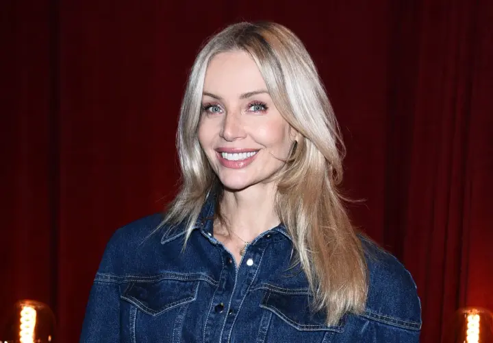 Uśmiechnięta blondynka z francuską grzywką w dżinsowej kurtce na tle czerwonej kurtyny.