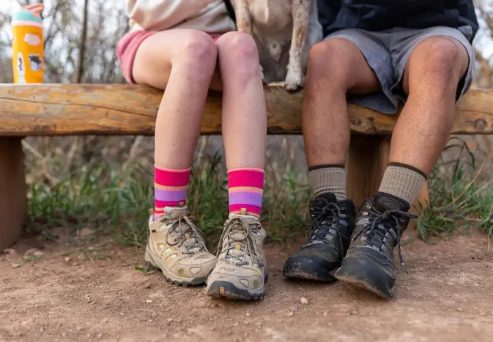 Colorful striped and neutral socks to wear with hiking boots, worn by two people sitting on a wooden bench with hiking boots on.
