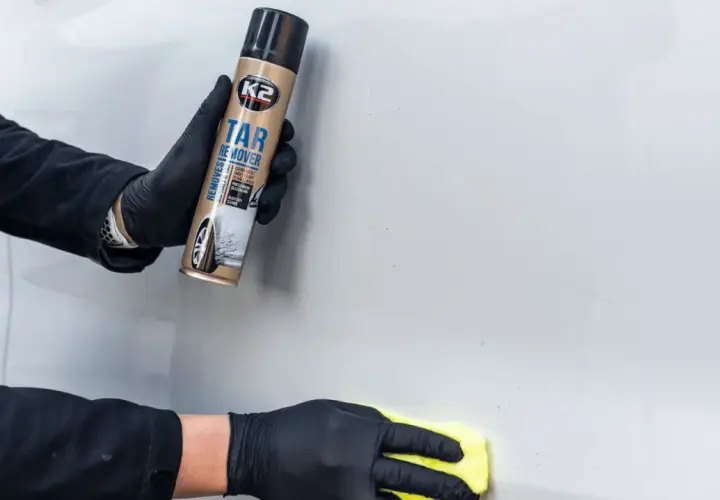 Ręce w czarnych rękawiczkach używają preparatu K2 Tar Remover, by usunąć klej z szyby auta.