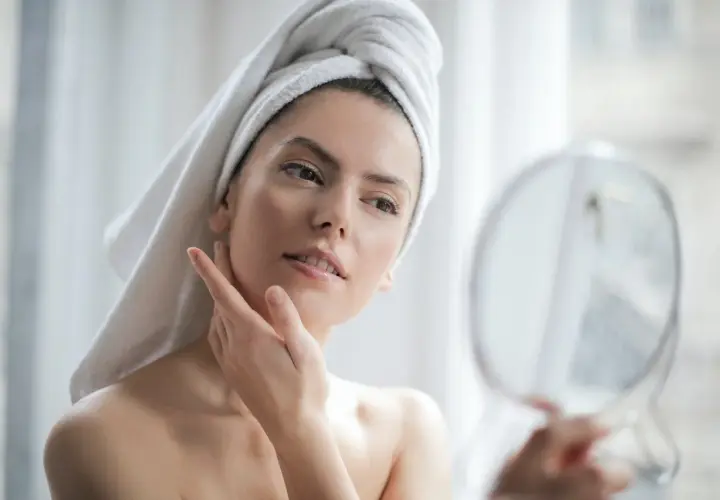 Donna con asciugamano in testa si guarda allo specchio, la sua pelle luminosa grazie all'acido ialuronico e vitamina C insieme.