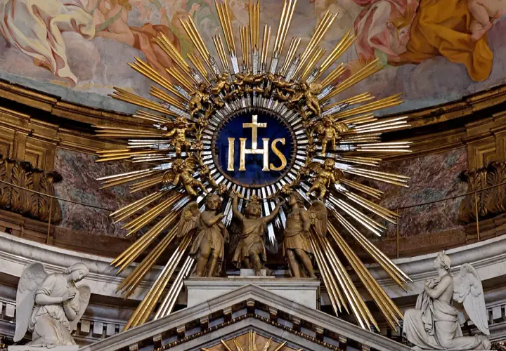 Goldene Strahlenkrone mit IHS-Symbol, das für Jesus steht. Putten und Engel umgeben das Symbol, das die göttliche Bedeutung von IHS unterstreicht.