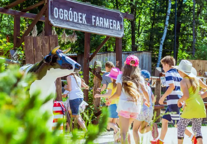 Dzieci biegną obok drewnianego szyldu "Ogródek Farmera" i figury krowy.