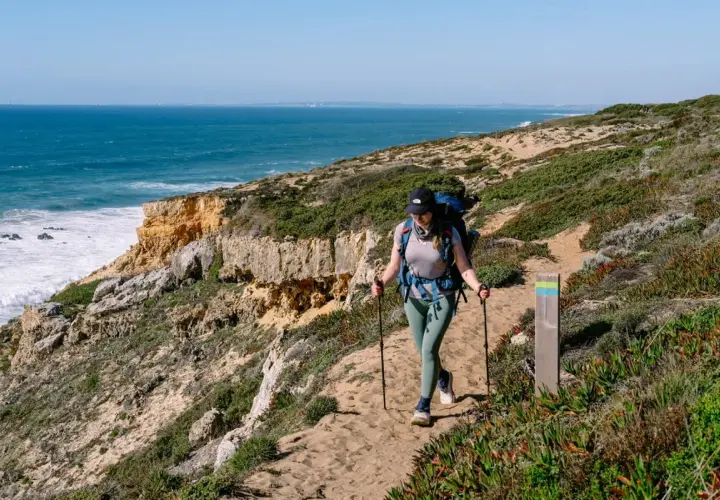 Wanderin auf dem Fisherman's Trail in Portugal. Eine Frau mit Rucksack und Stöcken wandert auf einem sandigen Küstenpfad mit Blick auf das Meer.