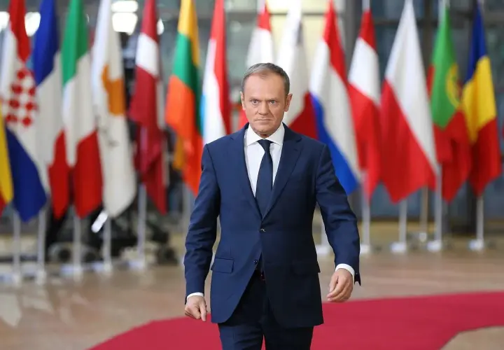 Donald Tusk, w garniturze, idzie na tle flag państwowych. Czy Donald Tusk jest Polakiem? Tak, jest.
