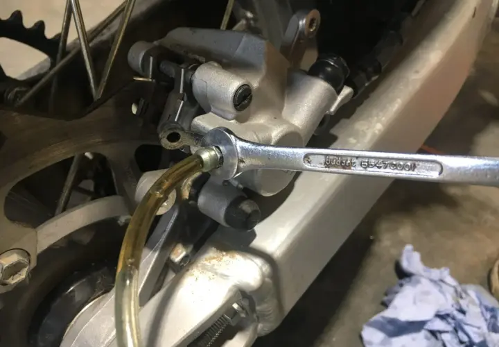 Klucz nasadowy odkręca odpowietrznik podczas wymiany płynu hamulcowego w motocyklu. Widać tarczę hamulcową i zacisk.