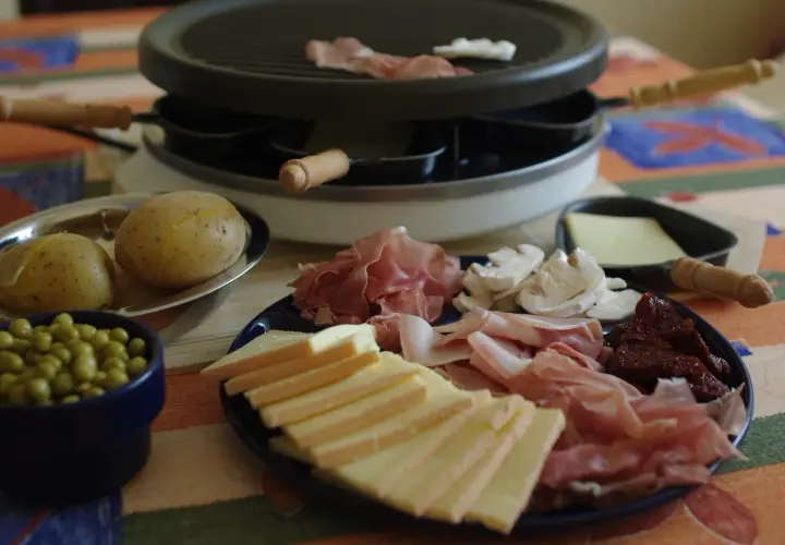 Préparation d'une raclette conviviale : fromage, charcuterie, pommes de terre et petits pois attendent sur la table.